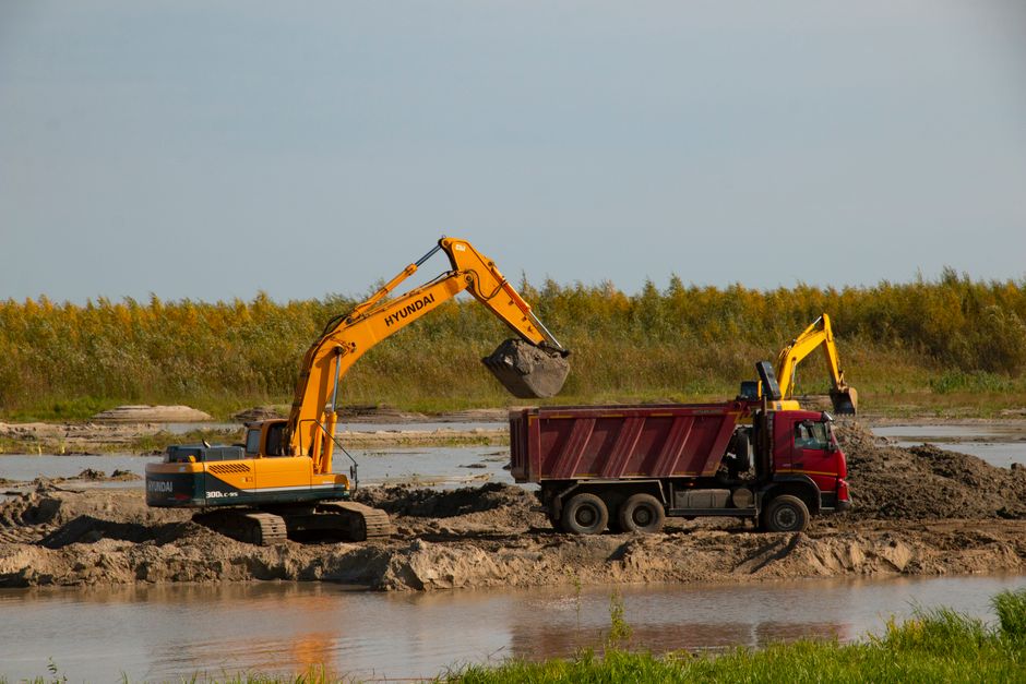Excavator transfers soil to a dump truck by a river at a construction site. Machinery in action.